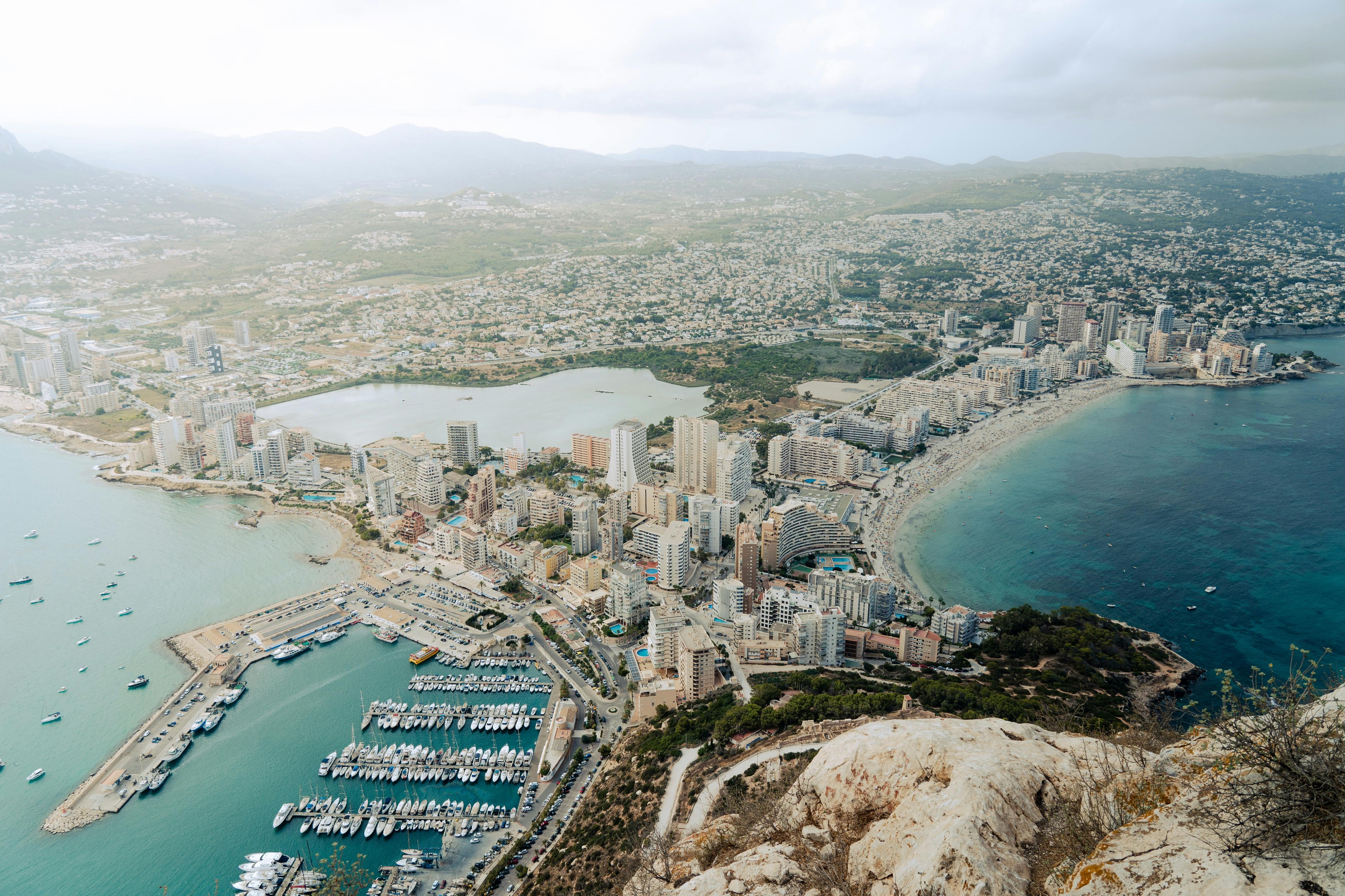 Vue panoramique de Calpe depuis le sommet du Peñón de Ifach, montrant le port de plaisance, les plages et les immeubles en bord de mer, avec les montagnes en arrière-plan.